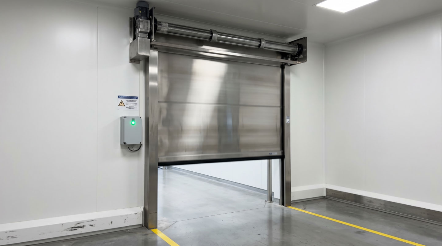 Porte industrielle rapide à enroulement en tablier inox fermée, encadrement métallique avec joints d'étanchéité visibles, dans un couloir de production aux murs blancs